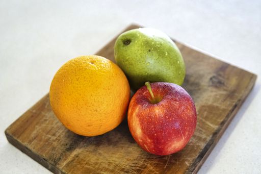 A closeup shot of a whole orange a green pear and a red  apple on a wooden cutting board