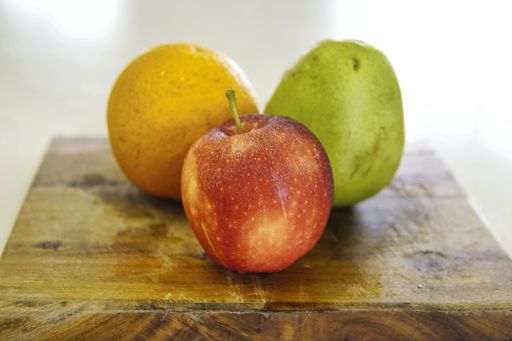 A closeup shot of a whole orange a green pear and a red  apple on a wooden cutting board