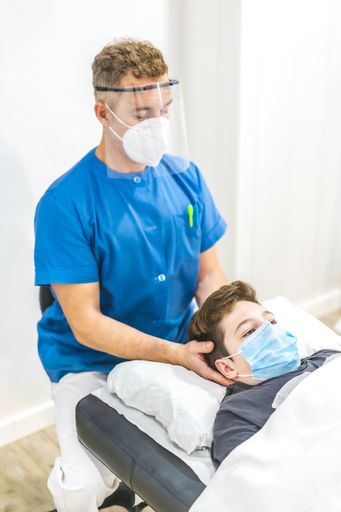 A vertical shot of a physiotherapist in a face mask giving neck Chiromassage to a young male patient