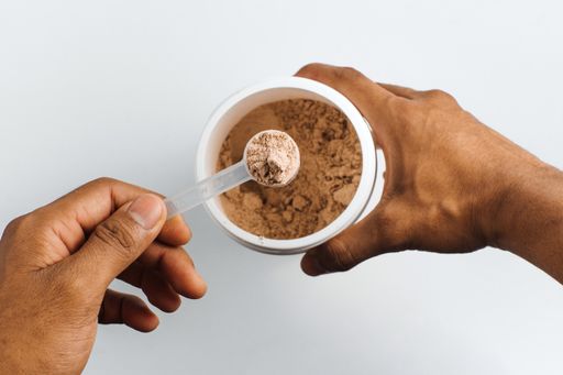 A person's hand holding a measuring spoon with chocolate protein powder over white background