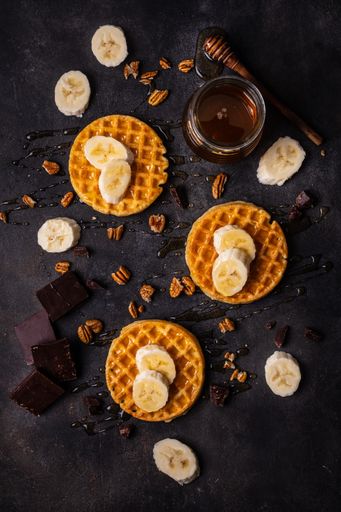 A vertical high angle shot of waffles with honey and bananas, and dark chocolate on a black surface