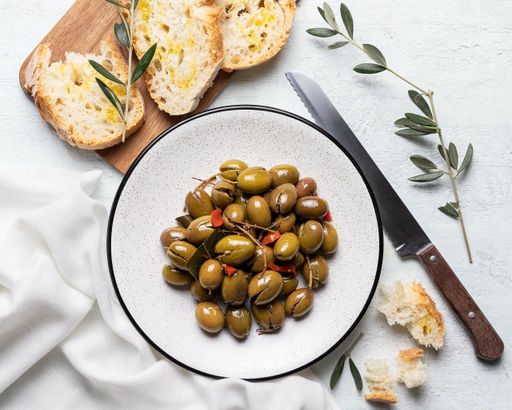 A top view of pickled green olives on a white plate with fresh bread and a knife on a white background