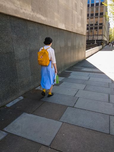 A young girl in a white shirt and long blue skirt walks alone down a London street carrying a yellow rucksack and a shopping bag.