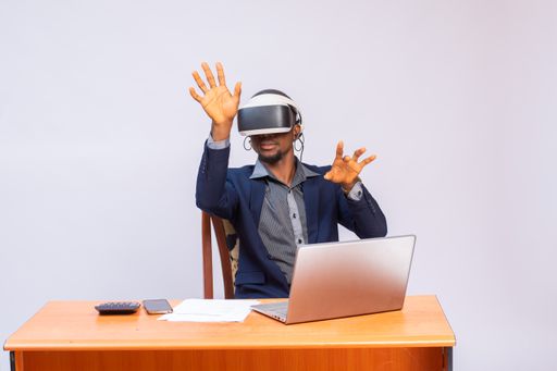 A handsome African man in a costume using VR glasses sitting next to a table with a laptop on it.