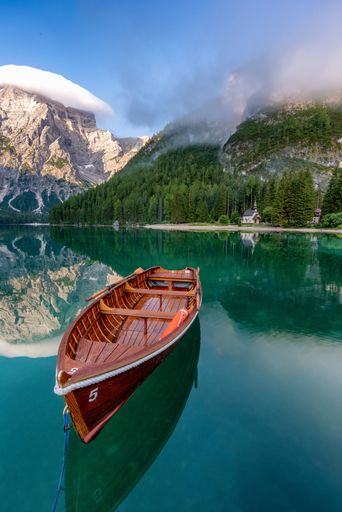 A vertical shot of an empty rowboat swimming in the lake in the background of cloudy mountains.