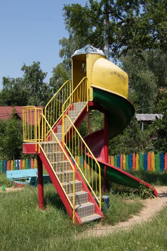 A vertical shot of a colorful playground slide with high trees and painted fence in the background