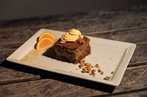 A horizontal shot of a piece of cake decorated with crushed walnuts, ice cream, and sliced oranges  on a white plate on a wooden surface