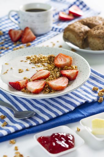 The delicious yogurt topped with sliced strawberries on a plate on a patterned fabric next to a spoon and some red jam in a plate