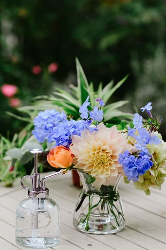 A vertical shot of a different kinds of colorful flowers in a pot with water on the table and a beautiful green garden in the background