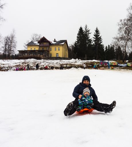 A woman and toddler boy sliding with a red sled on snow during a cold winter day at a park