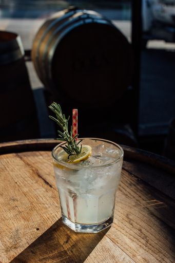 A vertical closeup of a rosemary lemonade soda with ice and a picnic gingham drinking straw on a whiskey barrel