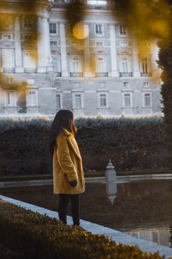 A vertical selective shot of a female wearing yellow coat standing by the water near a white building