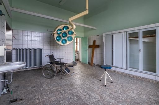 A wheelchair in an abandoned and creepy hospital. In the background an old grave cross in the operating room.