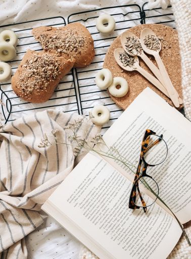 An overhead shot of an opened book with glasses on top of it, baked bread, flowers and towels