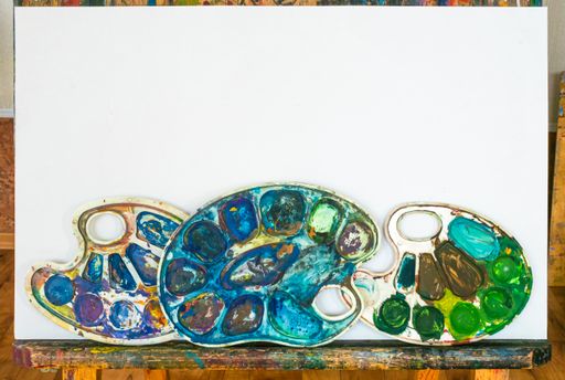 Blank sheet of white paper stands on wooden art easel. Below are three dirty color palettes. Workplace of the artist.