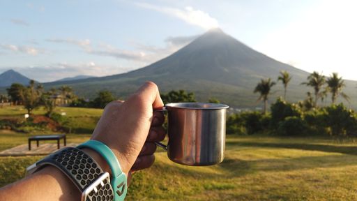 A person's hand holding a metal cup against a beautiful view of the Mayon volcano in Legazpi, Philippines