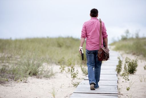 A male walking on a wooden pathway carrying his bag and holding the bible with a blurred background