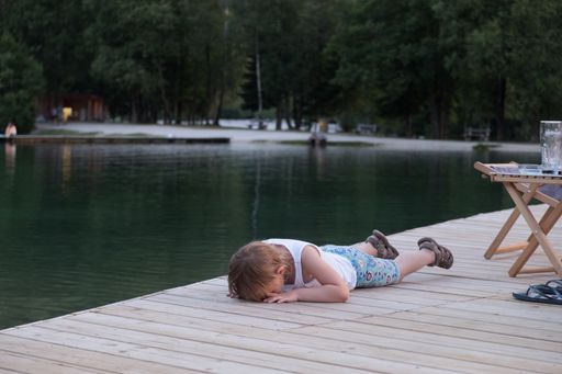 A beautiful view of a male kid sleeping on a wooden surface near a water lake with trees in the park