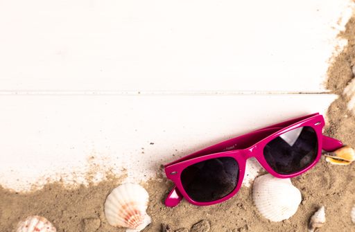 A top view of pink sunglasses with sand and shellfish on white wooden background