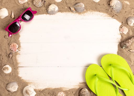 A top view of a frame with sunglasses, sand, shellfish and flip flops on white wooden background