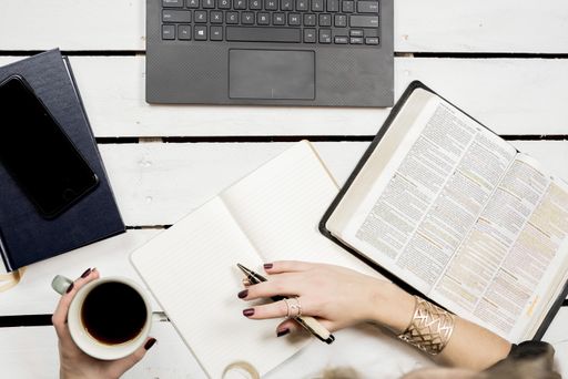 A overhead closeup shot of a person working at a table with a book, notebook, laptop, phone and a cup of coffee
