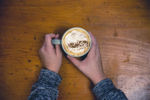 An overhead selective shot of a person holding a ceramic cup with latte art coffee on a wooden surface