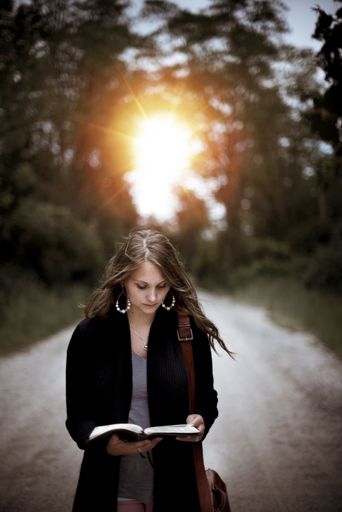 A vertical shot of a female standing on an empty road while reading the bible with the sun in the background