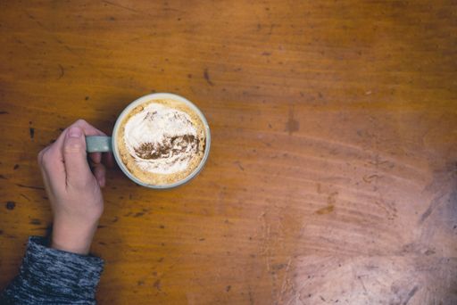An overhead selective shot of a person holding a ceramic cup with latte art coffee on a wooden surface