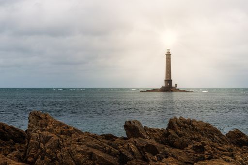 A  scenic view of a lighthouse built on water near the shore with the sun shining brightly on top of it