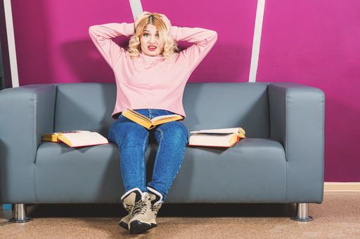 Young blonde girl sits on sofa in room and holds head with hands. On face of emotion of panic. Around her are open books. Difficulties in learning.