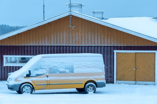 A yellow Ford Transit van covered in snow in front of a colored door of barn in Finland