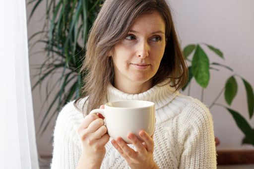 A charming woman with a white sweater drinking a mug of tea while sitting at home and enjoying the leisure time of the day