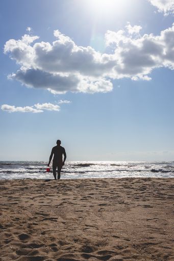 A vertical shot of a silhouette of a young man with a sand bucket in a sandy beach approaching camera
