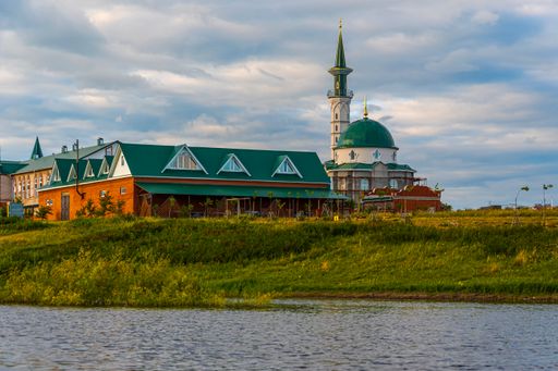 Complex of Muslim buildings with mosque, minaret and madrasah on banks of river against blue cloudy summer sky.