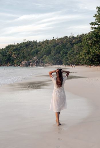 A vertical view of the Caucasian female walking on the sandy beach with white dress in Seychelles