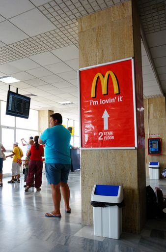 A vertical shot of people standing in an airport waiting room next to a column with a Mcdonald's sign.