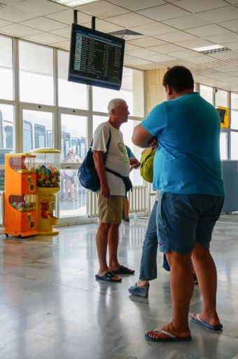 A vertical shot of people waiting next to a screen with departure schedules inside an airport.