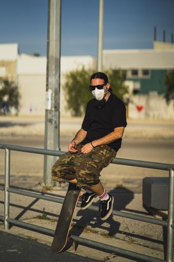 A vertical shot of a male wearing a face mask sitting on metal railings with a skateboard below- Covid-19 concept