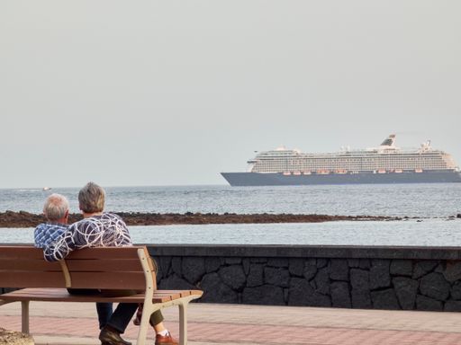 An elderly couple sitting on a bench overlooking the sea and a cruise ship in the background