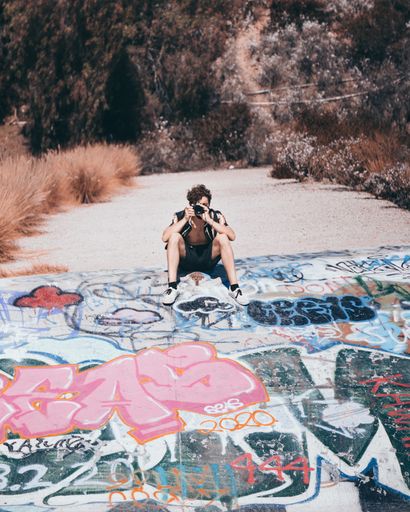 A vertical shot of a male with a camera sitting on a graffiti ground on a sunny day