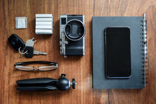 An old camera, batteries, tripod, memory card, car keys, eyeglasses, phone and a notebook on a table