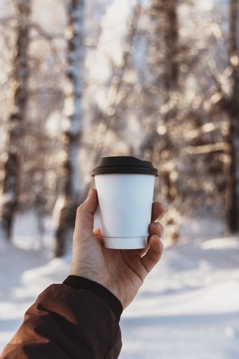 A vertical selective focus shot of a Caucasian man's hand holding a paper cup, with bare trees and snow in the background