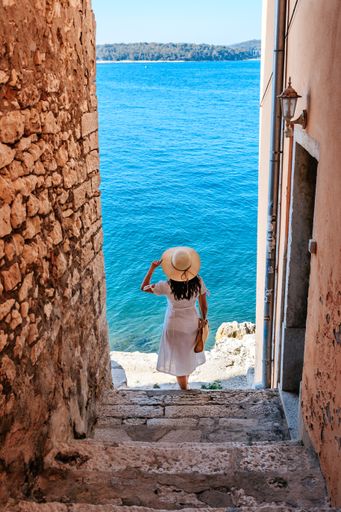 A vertical shot of a rear view of a young woman in a white dress standing in a narrow alley leading to the sea in Rovinj, Croatia