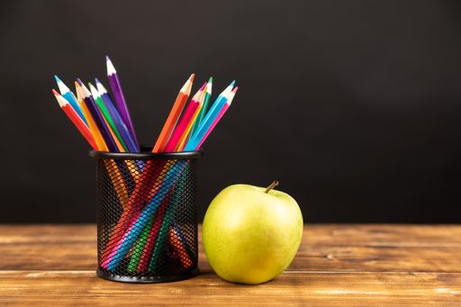 A stack of colored pencils with a green apple on wooden background with chalkboard school