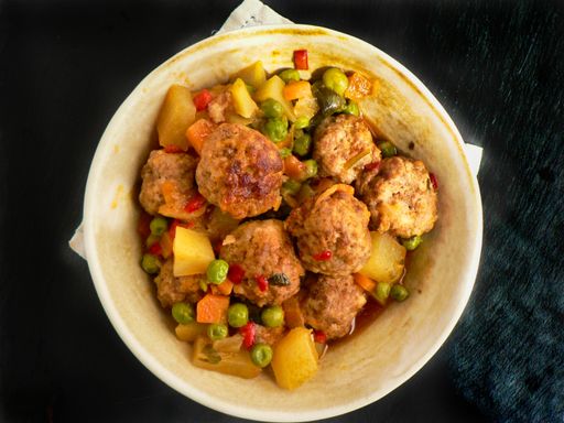 A top view of meatballs with potatoes and peas in a clay pot on a black surface