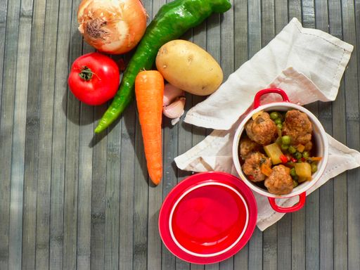 A top view of a pot of meatballs with potatoes and peas and raw vegetables on a wooden table