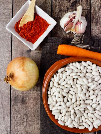 A top view of white beans in a clay bowl and other ingredients for beans stew - paprika, onions, garlic carrots on a wooden table
