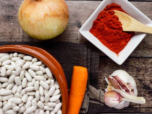 A top view of white beans in a clay bowl and other ingredients for beans stew - paprika, onions, garlic carrots on a wooden table