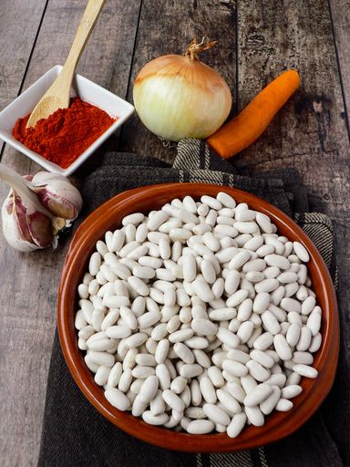 A top view of white beans in a clay bowl and other ingredients for beans stew - paprika, onions, garlic carrots on a wooden table