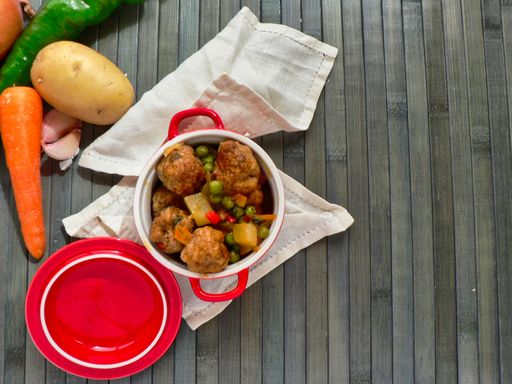 A top view of a pot of meatballs with potatoes and peas and raw vegetables on a wooden table